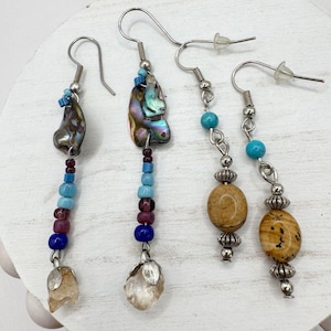 May include: Four pairs of dangle earrings with unique designs. Two pairs feature iridescent abalone shells and colorful beads. The other two pairs have turquoise beads and oval-shaped stones. All earrings have silver-toned hooks.