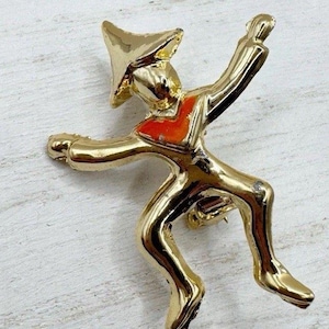 Könnte beinhalten: Eine goldfarbene Metallbrosche in Form einer stilisierten Figur mit Hut und roter Weste. Die Figur befindet sich in einer dynamischen Pose, möglicherweise tanzend oder springend, mit ausgestreckten Armen. Die Brosche hat eine glänzende Oberfläche.