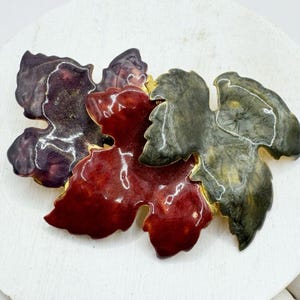 Puede incluir: Broche vintage con un grupo de hojas estilizadas en tonos burdeos, rojo y verde oliva. Las hojas tienen un acabado brillante con detalles dorados. El broche probablemente está hecho de metal y está diseñado para ser usado como un accesorio decorativo.