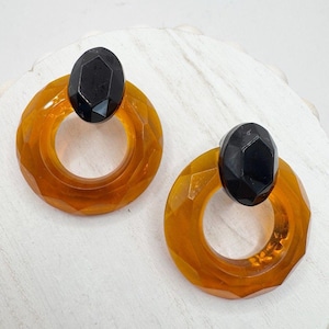 May include: A pair of amber-colored, geometric hoop earrings with a black, oval-shaped accent. The earrings have a faceted design, creating a play of light and shadow. These vintage-inspired earrings are a bold statement piece.