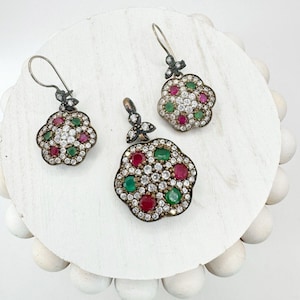 May include: A jewellery set comprising a pendant and two earrings. Each piece features a flower-shaped design with red, green, and clear gemstones set in a dark metal. The earrings have a hook closure.
