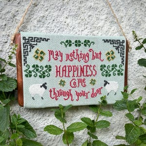 May include: A cross-stitched sign with the words "May nothing but HAPPINESS come through your door." The sign features a light green background, red and green lettering, and two white sheep. The sign is framed with wood and has a jute rope for hanging.