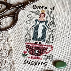 Può includere: Ricamo a punto croce di una donna in piedi su una tazza da tè, circondata da forbici. Il testo recita "Queen of Scissors".