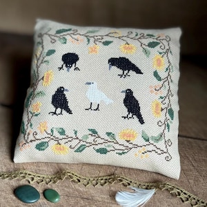 May include: A square, beige cross-stitch pillow featuring five black crows and one white bird. The birds are surrounded by a floral border of yellow flowers and green vines. A decorative gold lace trim and two green stones are in the foreground.