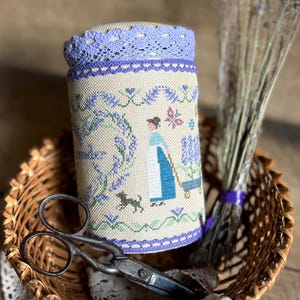 Op de afbeelding: Een cilindrisch speldenkussen met een geborduurd lavendelontwerp, met een vrouw die een kruiwagen duwt. Het kussen ligt in een rieten mand met een vintage schaar, kanten rand en een bos gedroogde lavendel. Het kussen heeft een lavendel kanten rand.