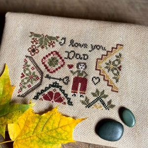 May include: A cross-stitch sampler with the text "I love you Dad" and a folk art design in red, green, and yellow. Two green stones and two yellow and green maple leaves are on a wooden surface.