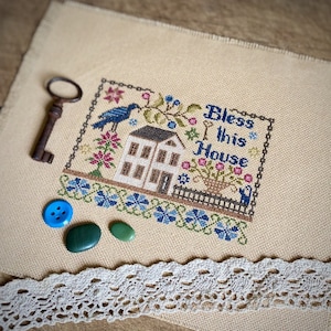 Peut inclure: Un motif de point de croix représentant une maison avec le texte "Bless this House" dans un cadre brun. La maison est blanche avec des bordures brunes et un oiseau bleu perché sur le toit. Le fond est un tissu de couleur crème.