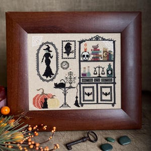 May include: A framed cross-stitch featuring a Halloween theme. The artwork includes a witch silhouette, a raven, a black cat, pumpkins, and a cabinet with potion bottles and a skull. The frame is made of dark brown wood.