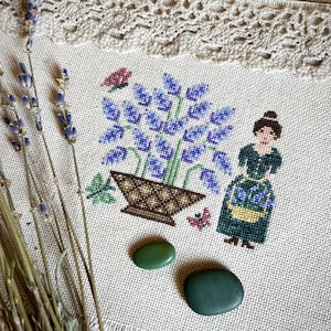 Puede incluir: Bordado de punto de cruz de una mujer sosteniendo una cesta de flores moradas, con una maceta de flores moradas en el fondo. El bordado está hecho en una tela de lino blanco con un borde marrón.