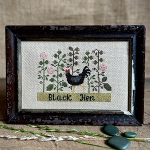 May include: Framed cross-stitch art featuring a black hen with a red comb, surrounded by pink flowers and green foliage. The text "Black Hen" is stitched below. The frame is dark brown with a distressed finish.