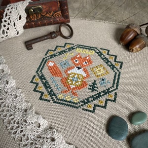 以下が含まれることがあります： 緑、黄色、オレンジの幾何学模様のフレームの中に狐が描かれたクロスステッチのパターン。パターンはベージュのリネン生地に刺繍されています。