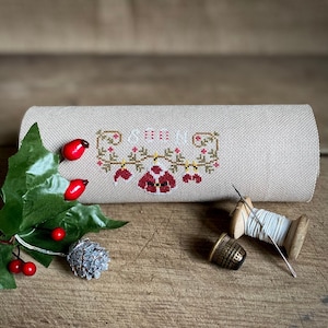 Può includere: Un tessuto di lino beige con un ricamo a punto croce che rappresenta una scena natalizia rossa e verde. Il tessuto è arrotolato e ci sono alcune bacche rosse, un ramoscello verde e una pigna argentata su una superficie di legno.