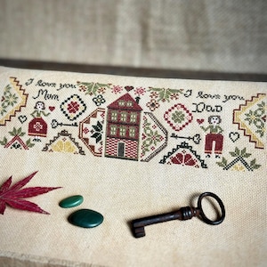 May include: Cross-stitch embroidery on a beige fabric with a house, two figures, and the text "I love you Mum" and "I love you Dad".