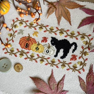 May include: A cross-stitch embroidery on light-colored fabric featuring three pumpkins with the letters B, O, and O stitched in black, accompanied by a black cat silhouette. The design is framed by a border of stitched acorns and autumn leaves.