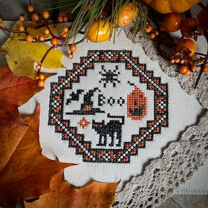 以下が含まれることがあります： 白地に黒と白の糸で刺繍されたクロスステッチ。魔女の帽子、クモ、カボチャ、黒い猫2匹がデザインされている。黒い糸で「BOO」と刺繍されている。黒と白の縁取りで囲まれている。