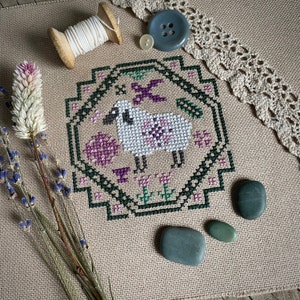 Peut inclure: Un motif de point de croix représentant un mouton blanc avec des fleurs roses et des feuilles vertes, encadré d'une bordure vert foncé. Le motif est sur un fond de tissu beige.