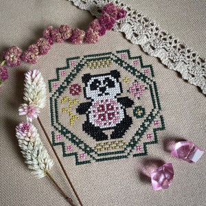 Puede incluir: Una bordado de punto de cruz de un oso panda con un diseño floral rosa y blanco en el centro. El panda está rodeado por un borde verde y amarillo con un patrón geométrico.