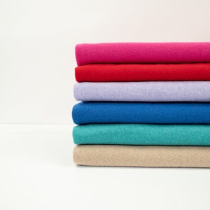 Könnte beinhalten: Ein Stapel gefalteter Stoffe in verschiedenen Farben: Pink, Rot, Lavendel, Blau, Türkis und Beige. Die Textilien sind ordentlich angeordnet und zeigen ein Farbspektrum. Das Bild ist vor einem schlichten weißen Hintergrund.