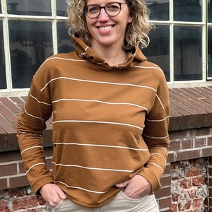 May include: A brown and white striped long-sleeved hooded sweatshirt with a relaxed fit. The sweatshirt is being worn by a person with curly blonde hair and glasses.