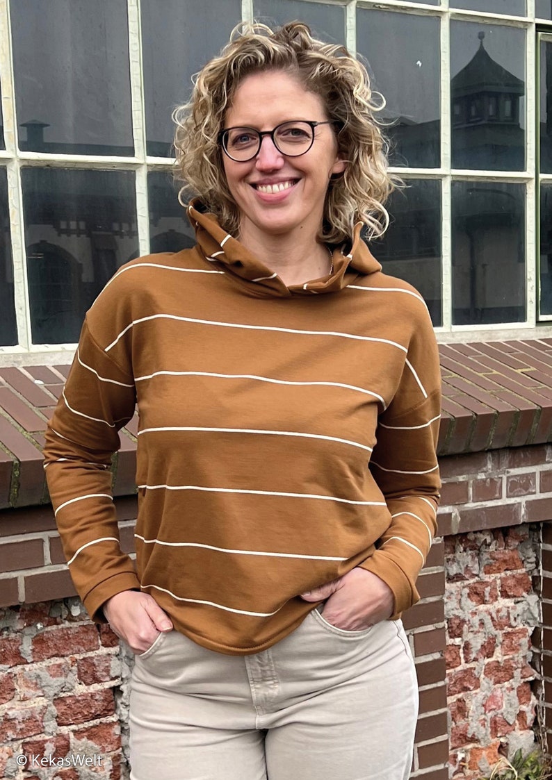 May include: A brown and white striped long-sleeved hooded sweatshirt with a relaxed fit. The sweatshirt is being worn by a person with curly blonde hair and glasses.
