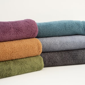 Pu&ograve; includere: Un mucchio di sei asciugamani in pile di diversi colori, tra cui viola, arancione, verde, grigio e blu. Gli asciugamani sono piegati e disposti ordinatamente su una superficie bianca.