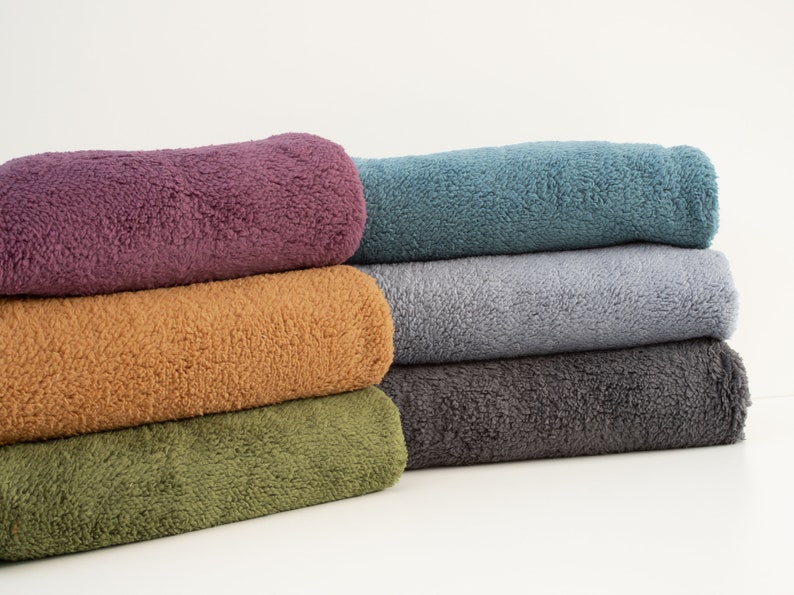 Pu&ograve; includere: Un mucchio di sei asciugamani in pile di diversi colori, tra cui viola, arancione, verde, grigio e blu. Gli asciugamani sono piegati e disposti ordinatamente su una superficie bianca.