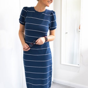 May include: A woman wearing a blue and white striped maxi dress with short sleeves. The dress has a fitted bodice and a flowy skirt. The woman is smiling and holding a pair of sunglasses.