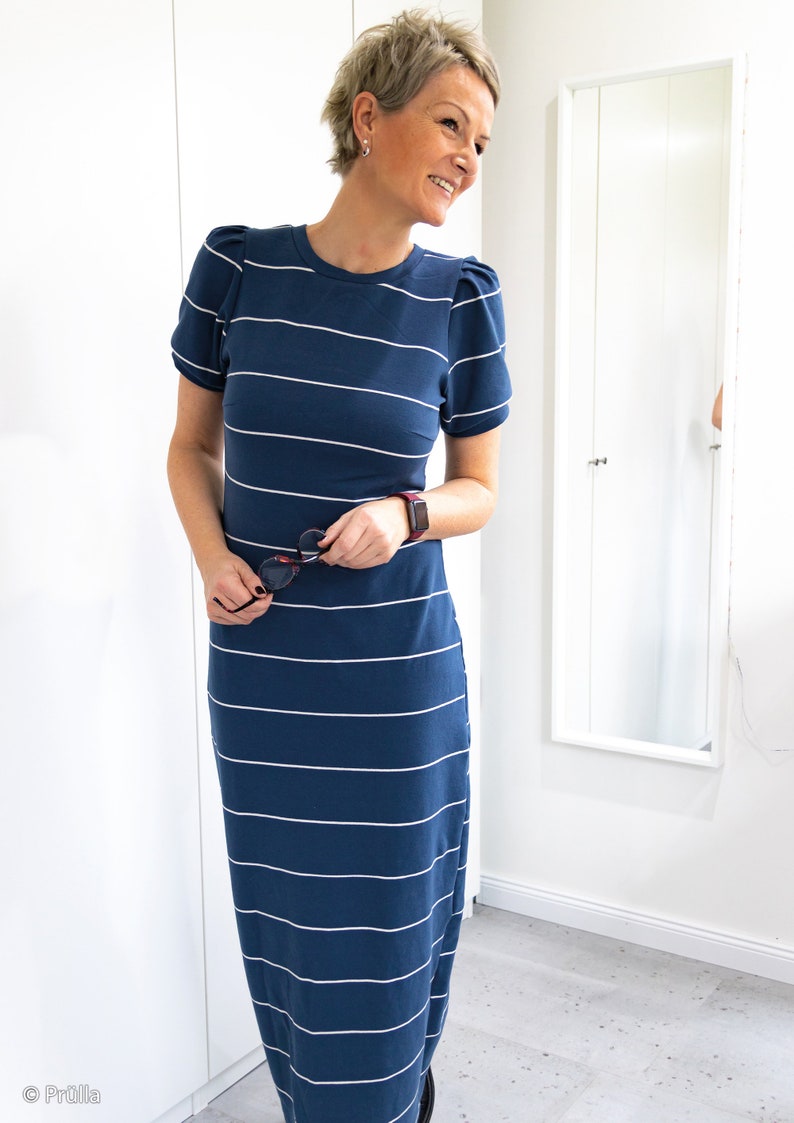 May include: A woman wearing a blue and white striped maxi dress with short sleeves. The dress has a fitted bodice and a flowy skirt. The woman is smiling and holding a pair of sunglasses.