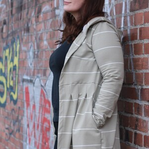 May include: A woman wearing a long, striped, hooded cardigan in a light beige colour with white stripes. She is standing in front of a brick wall with graffiti.