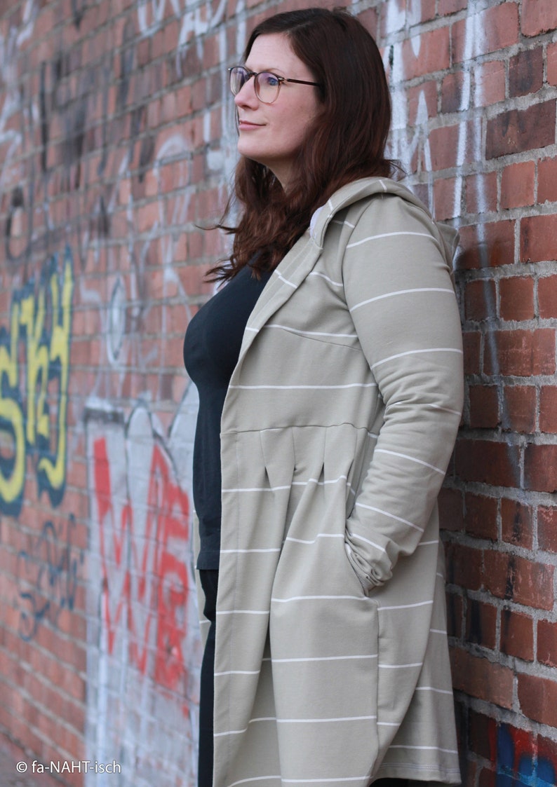May include: A woman wearing a long, striped, hooded cardigan in a light beige colour with white stripes. She is standing in front of a brick wall with graffiti.