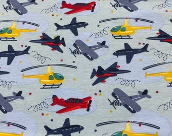 May include: A blue and white fabric with a repeating pattern of airplanes, helicopters, and stars. The airplanes are in various colors, including red, blue, and yellow. The helicopters are yellow and have a white rotor.