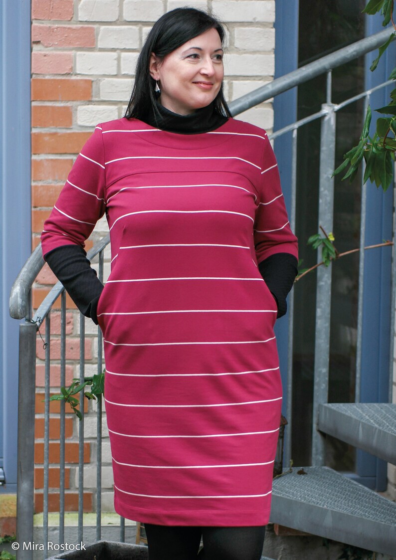 May include: A woman wearing a pink and white striped dress with long sleeves and black trim. The dress has a scoop neck and pockets on the sides.