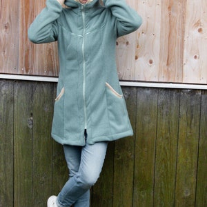 Pu&ograve; includere: Una donna indossa una giacca di pile color verde acqua con cerniera e due tasche. Indossa jeans blu e scarpe da ginnastica bianche.