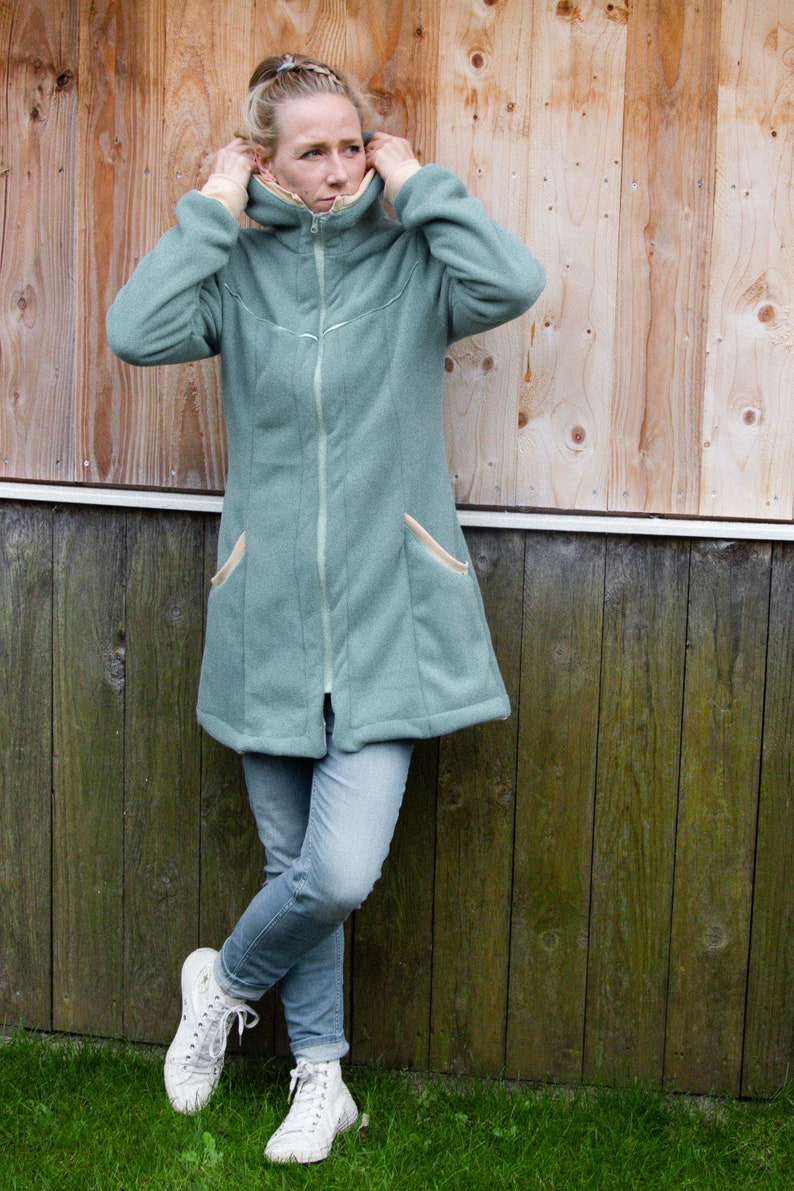 Pu&ograve; includere: Una donna indossa una giacca di pile color verde acqua con cerniera e due tasche. Indossa jeans blu e scarpe da ginnastica bianche.