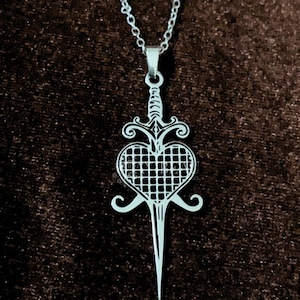 Könnte beinhalten: Silberfarbene Halskette mit Anhänger. Der Anhänger zeigt ein Schwertdesign mit einem herzförmigen Käfig in der Mitte. Der Griff ist verziert, und die Klinge ist lang und spitz. Die Kette ist ebenfalls silberfarben.