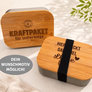 Könnte beinhalten: Zwei rechteckige Brotdosen mit hellbraunen Bambusdeckeln und grauen Unterteilen. Ein Deckel trägt den eingravierten Text KRAFTPAKET für unterwegs und ein kreisförmiges Logo. Der andere Deckel hat eingravierten Text und ein schwarzes Gummiband.