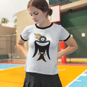 May include: White t-shirt with a black ringer and a graphic of a stylized black bird with a yellow beak and eye.