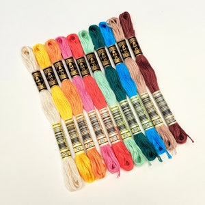 Könnte beinhalten: Eine Sammlung von 12 Stickgarnrollen in verschiedenen Farben, darunter Ecru, Gelb, Orange, Rosa, Grün, Blau und Braun. Jede Rolle hat ein goldenes Etikett mit dem DMC-Logo und einer darauf gedruckten Nummer. Die Nummern auf den Rollen sind: ECRU, 743, 722, 603, 3705, 955, 991, 3846, 3864 und 3858.