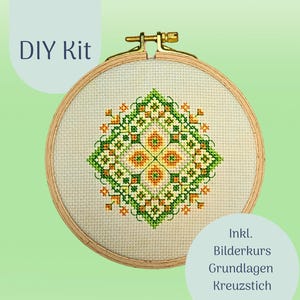Könnte beinhalten: Ein DIY-Kreuzstich-Set mit einem geometrischen Design in Grün, Orange und Gelb. Das Design befindet sich auf einem cremefarbenen Stoff, der in einem hölzernen Stickrahmen gespannt ist. Der Text "DIY Kit" ist sichtbar. Das Set enthält einen Bilderkurs und Grundanleitungen.