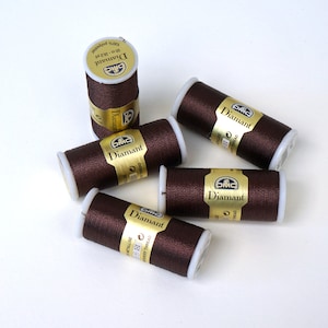 Könnte beinhalten: Fünf Spulen braunes Stickgarn mit goldenen Etiketten mit der Aufschrift "DMC Diamant 100% Polyester 50 m - 32/3"