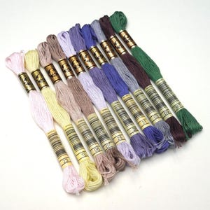 Peut inclure: Assortiment de 12 bobines de fil à broder dans différentes nuances de rose, violet, bleu, vert, marron et beige. Chaque bobine porte une étiquette dorée avec un numéro imprimé dessus.
