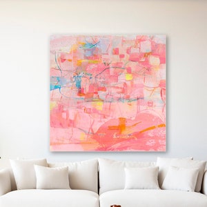May include: Abstract painting with pink, orange, yellow, and blue colors. The painting is on a white canvas and is hanging on a white wall in a living room setting. A white couch, a wooden coffee table, and a plant in a woven basket are in the foreground.