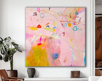 Op de afbeelding: Abstract schilderij met roze, gele, blauwe en groene kleuren. Het schilderij hangt aan een witte muur in een woonkamer met een witte bank, twee stoelen en een plant.