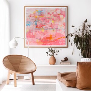 May include: Abstract painting in shades of pink, yellow, and blue, framed in a natural wood frame, hanging on a white wall above a wicker chair and a brown leather couch.