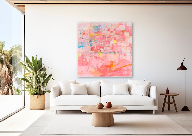 May include: Abstract painting with pink, orange, yellow, and blue colors. The painting is on a white canvas and is hanging on a white wall in a living room setting. A white couch, a wooden coffee table, and a plant in a woven basket are in the foreground.