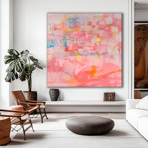May include: Abstract painting with pink, orange, yellow, and blue hues. The painting is framed and hanging on a white wall in a living room setting.