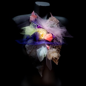Può includere: Un arrangiamento di fiori in tessuto colorato con strati di tulle, pizzo e nastro. L'arrangiamento è in tonalità di rosa, giallo, viola e verde.