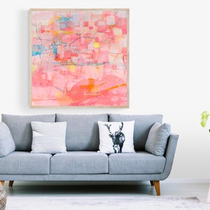 May include: A gray sofa with three pillows, one with a deer design, in a living room with a framed abstract painting in pink and yellow hues, a potted plant on a white stool, and a woven basket on the floor.