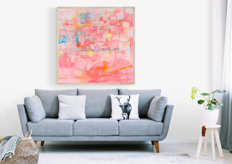 May include: A gray sofa with three pillows, one with a deer design, in a living room with a framed abstract painting in pink and yellow hues, a potted plant on a white stool, and a woven basket on the floor.