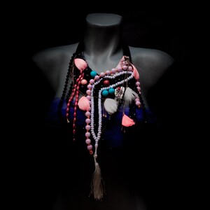 Può includere: Una collana a strati con perline rosa, blu e bianche e un ciondolo d'argento. La collana è su un manichino e ha un fiocco che pende dal fondo.